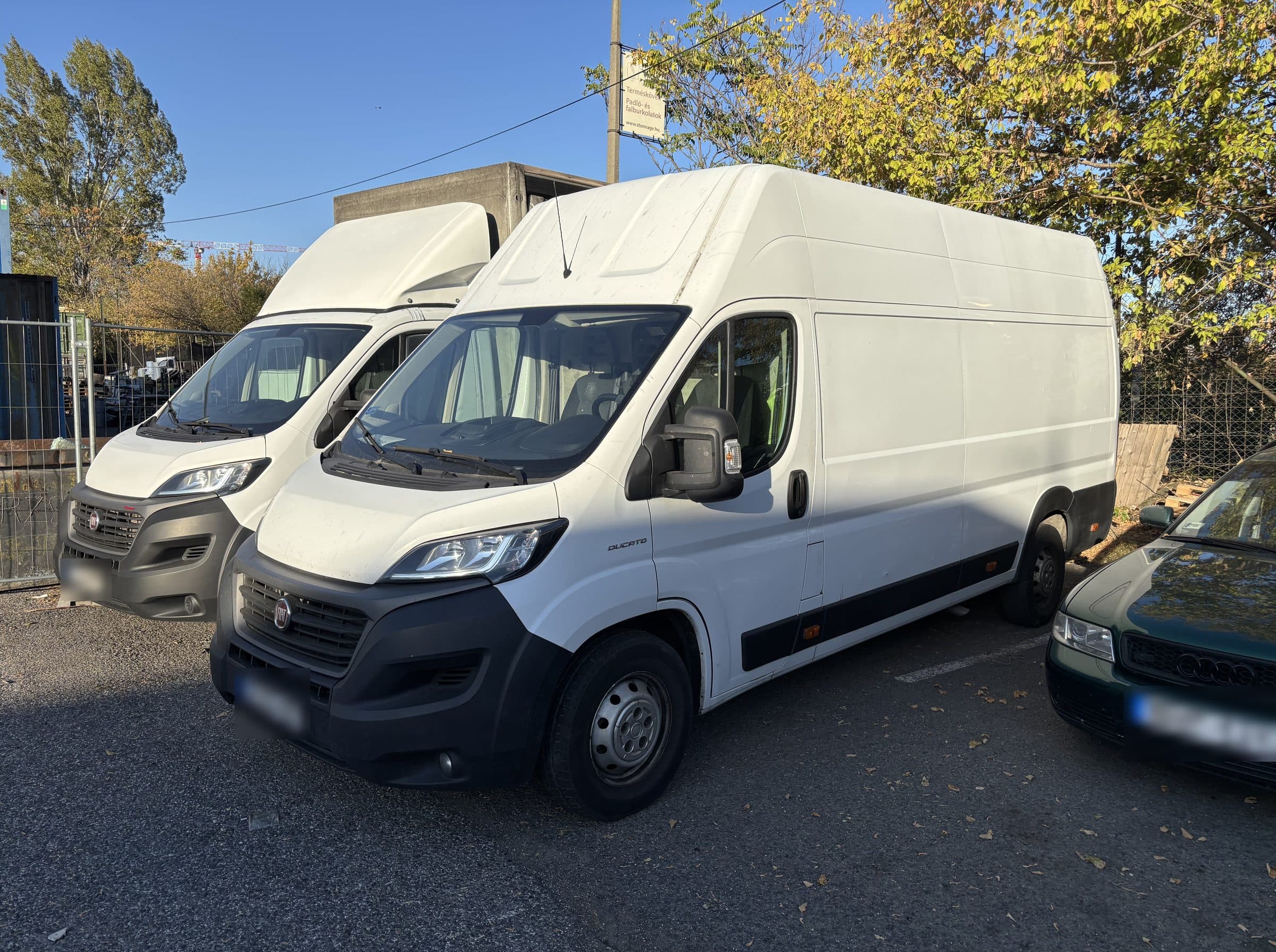 Fehér, magas tetős Fiat Ducato Maxi zárt dobozos furgon elölről, mögötte egy másik Ducato, napsütéses telephelyen